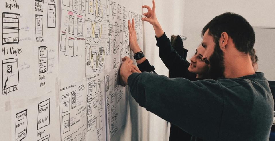 Two people collaborating on hand-drawn UI wireframes taped to a wall