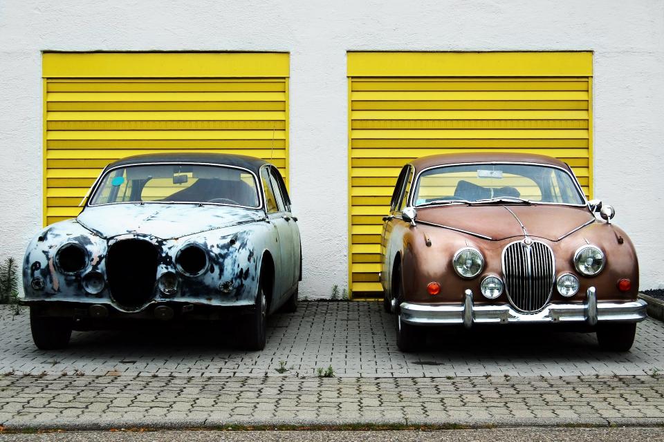 Two vintage cars side-by-side. The one on the left is rusted out and missing headlights. The one on the right has been restored to good condition