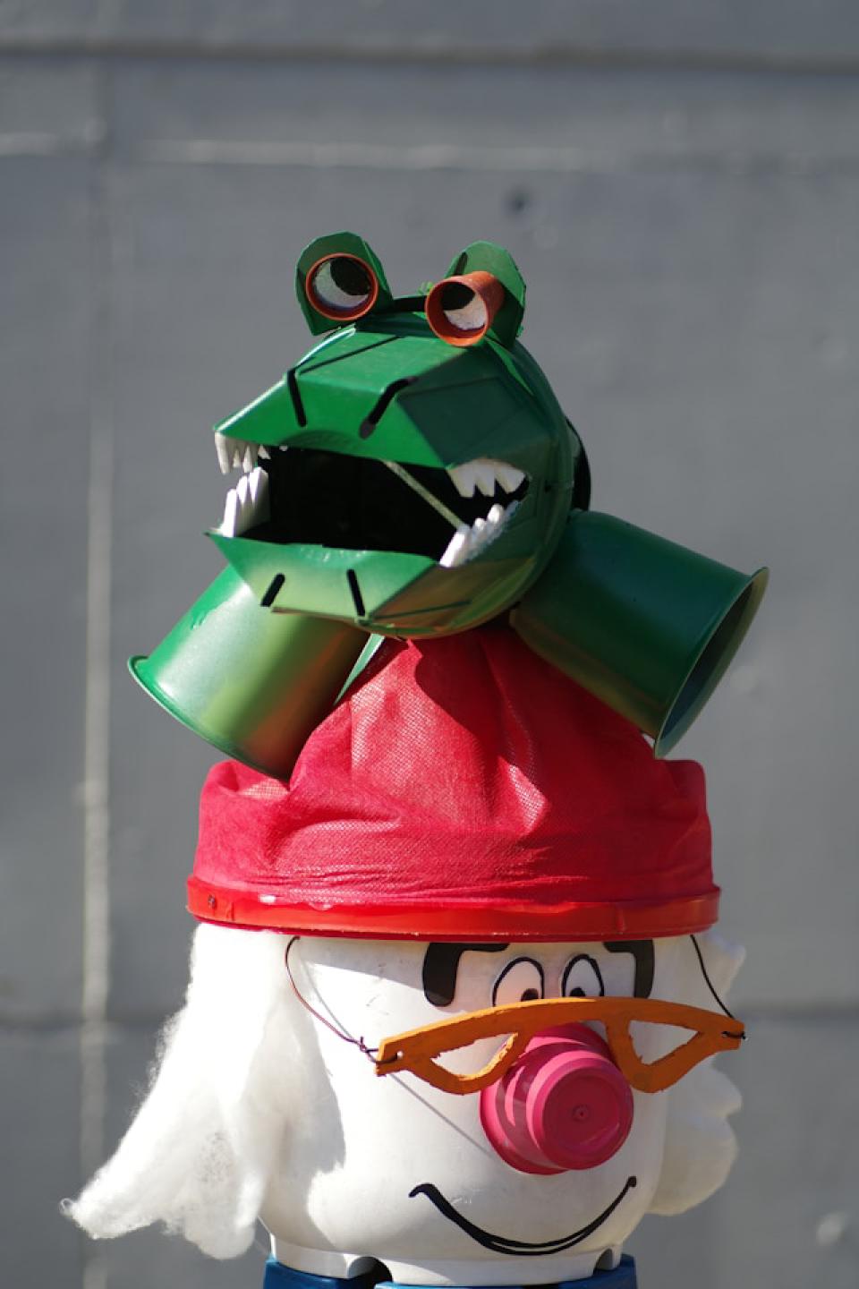 A funky crocodile made out of cups and other materials. It is sitting on top of some other cartoon character, wearing a read hat